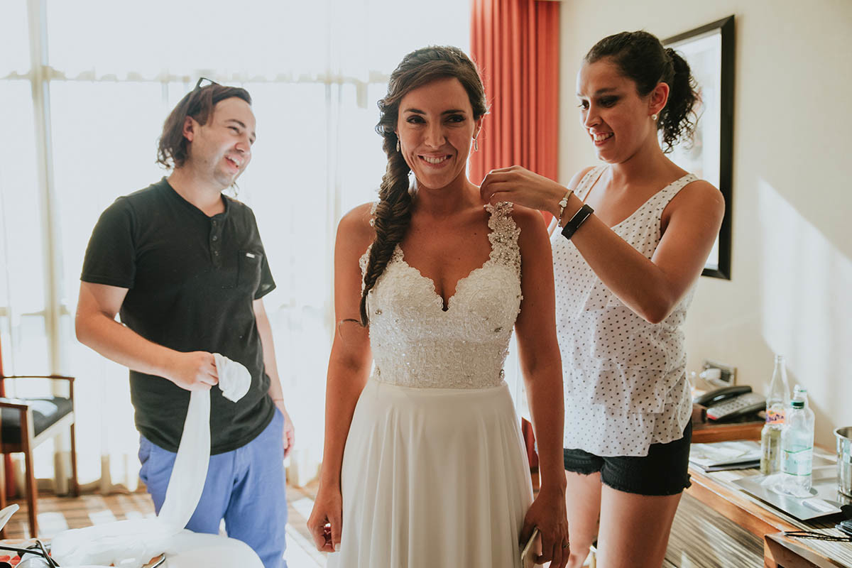 una mujer con vestido de chiffon corte en a blanco una mujer costurera ajustando el vestido un hombre con camisa negra y pantalón jeans en habitación preparando una novia para el casamiento