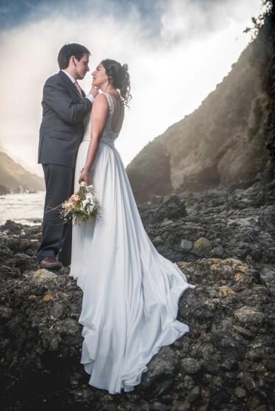 Novios besando al aire libre sobre rocas frente al mar en el sur de Chile el novio viste un terno marengo y corbata y la novia lleva puesto un vestido con escote en la espalda, cola en chiffon y tiene una rama de flores en la mano